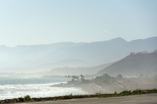 An unpopulated two-lane highway stretches out to the horizon with majestic mountains and a stunning ocean view in the background
