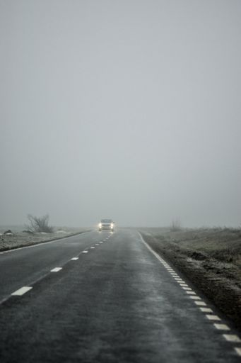 A vertical shot of a car driving along the asphalt road through the countryside on a foggy day
