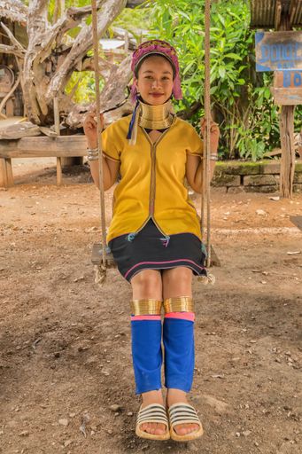 Karen woman from Pai at Mae Hong Son, Thailand, with brass neck rings, sits on wooden swing in rustic village, wearing vibrant color Karen dress