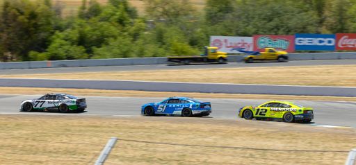 Two yellow race cars speeding around a motor racing track, with other vehicles parked in the background