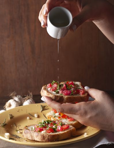 A vertical shot of a woman holding a sandwich with tomato and rosemary while pouring oil onto it