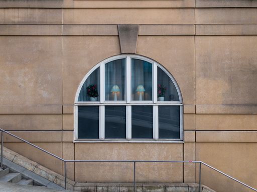 A window on a stone building with lamps and vases in the background seen through urban stairs