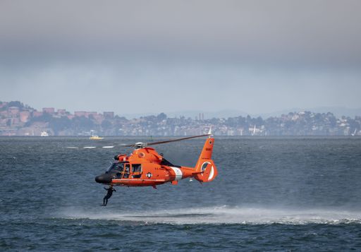 Aerial view of a vibrant orange helicopter flying over a vast blue body of water, with a sprawling cityscape in the background
