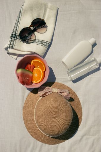 A vertical shot of assorted fruits in a pink bowl, a hat, sunglasses, sun cream on a white fabric