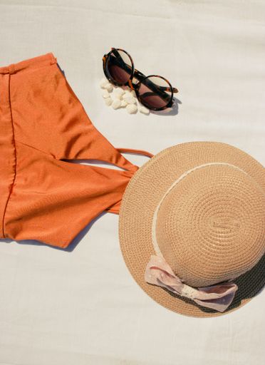 A vertical shot of orange bikini, a hat, and sunglasses on a white fabric background