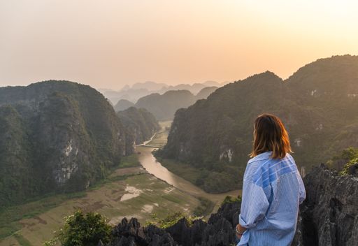 An adult female stands in a picturesque landscape, looking out over a river snaking through the mountain range in the background