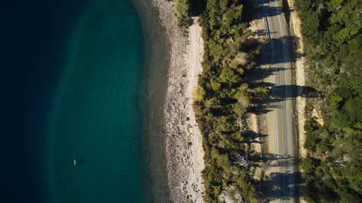 Birds eye view of a road in a forest with big trees and a lake shore with turquoise waters. Sunset in Villa la Angostura, patagonia Argentina.