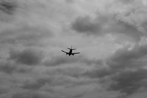 A grayscale shot of the silhouette of a plane flying under vast gray and white clouds in the sky