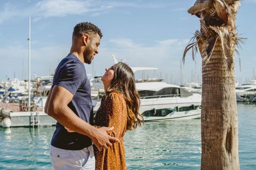 Picture of Couple Cuddling and Laughing at the Port with Boats Floating in Water Behind and Palm Tree (Black Boyfriend and Hispanic Girl looking at ea