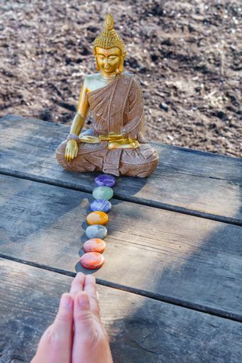 woman's hands meditating in front of a Buddha statue with colorful chakra stones on a wooden table in a park in the sunshine