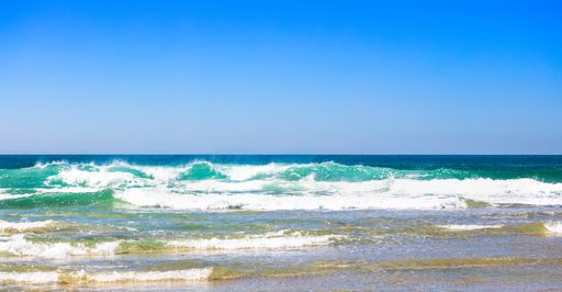 A panoramic shot of foamy ocean waves breaking on a sandy beach under a blue cloudless sky