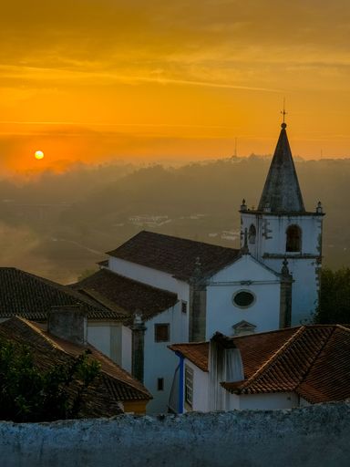 A stunning outdoor shot of a white church in the foreground with a backdrop of mountains and trees illuminated in a warm, orange sunset