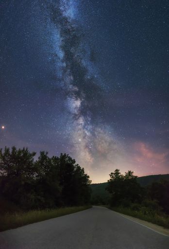 A breathtaking scenery of an empty road surrounded by beautiful green trees under the Milky Way Galaxy