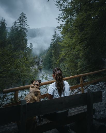 A female sitting on a wooden bench with her pet dog in a wooded area.