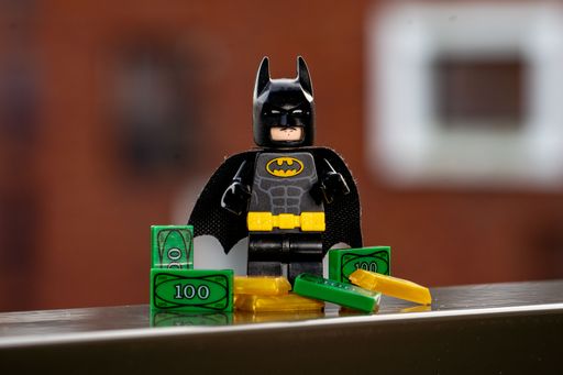 A Lego mini figure of Batman standing behind a pile of money and gold.