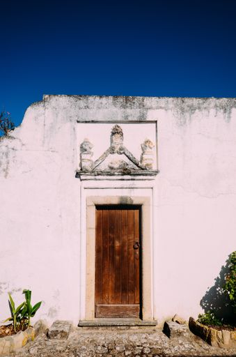 A vertical shot of an old, pink building with a wooden door and ornament under a blue sky in Obidos