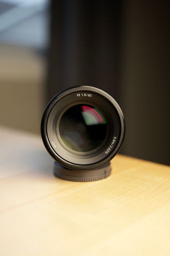 A DSLR camera lens is nestled on a wooden table beside a laptop computer, with a blurred background and natural lighting