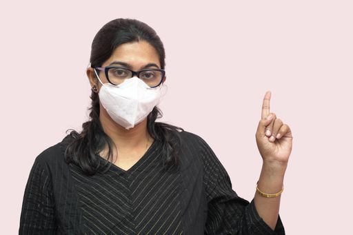 A young woman from India in a facemask pointing her finger up with a pink background