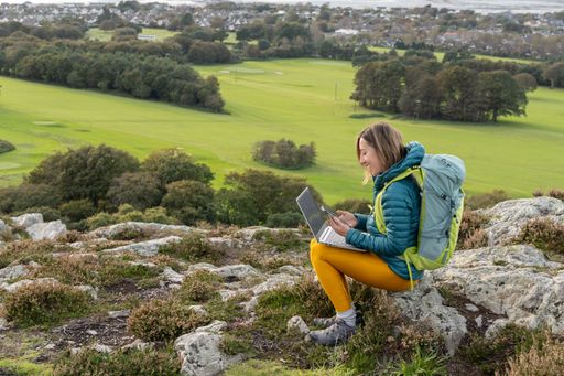 A young adult woman in her twenties wearing a blue outdoor coat, using a laptop computer while sitting outdoors in a mountainous region