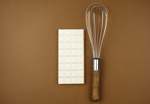 A top view of a white milk chocolate bar next to a wire whisk on a soft brown background