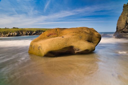 A rock on the beach surrounded by the sea with long exposure and cliffs under a blue sky