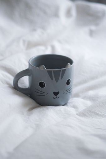 A mug with a black cat face design is lying flat on a white bedspread