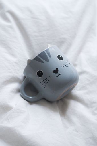 A mug with a black cat face design is lying flat on a white bedspread