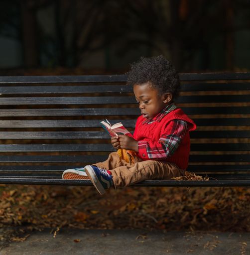 An adorable young African child sitting on a bench in a park and reading from a notebook