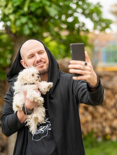 A young Hispanic male wearing a black jacket with a cute white dog taking a selfie