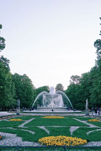 An outdoor scene featuring a fountain surrounded by a variety of colorful flowers, all enclosed in a decorative white picket fence