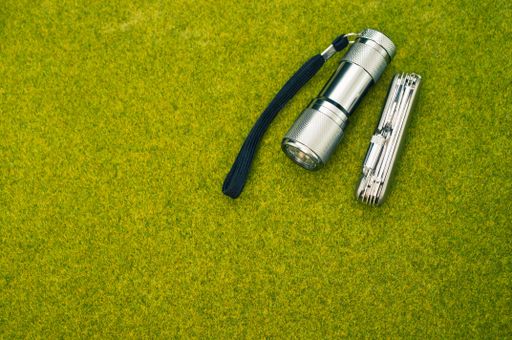 A top view shot of a steel knife and a flashlight on a green grass surface