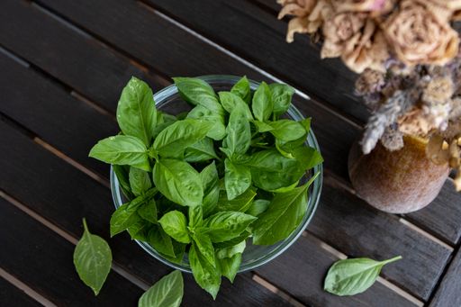 A bowl full of fresh basil is on the wooden table beside of a vase of preserved flowers.