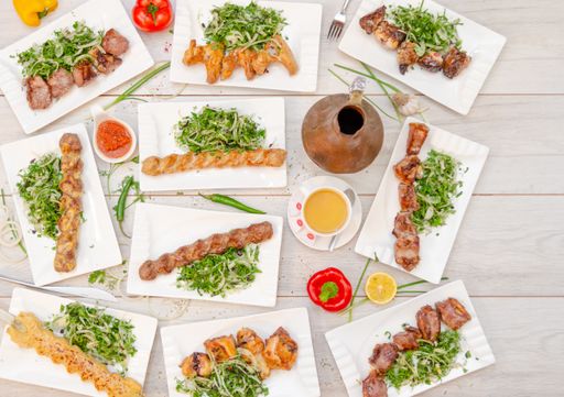 A flat lay of barbecued kebabs with hot peppers, spicy sauces, and a clay pot of tea in the middle