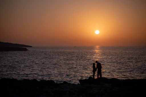 Two individuals standing on rocky shore by the ocean against the sun