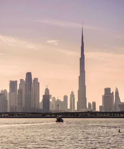 A vertical shot of a boat travelling across the sea with city buildings and a beautiful scenery of sunset in the background