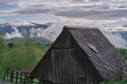 A small wooden building in a village surrounded by hills covered in greenery and fog under a cloudy sky