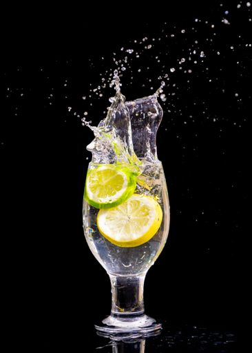A vertical shot of  lemon slices splashing into glass water on isolated black background