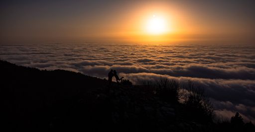 Two people stand on top of a mountain, enjoying an amazing view of the clouds below them
