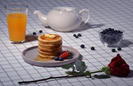 A white ceramic plate topped with three golden-brown pancakes, adorned with a light rose flower next to a tall glass of freshly-squeezed orange juice