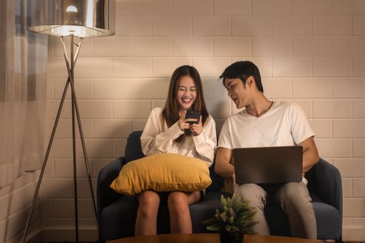 Young couple relaxing on couch with devices, woman laughs at phone content and sharing a funny moment together, cozy home setting