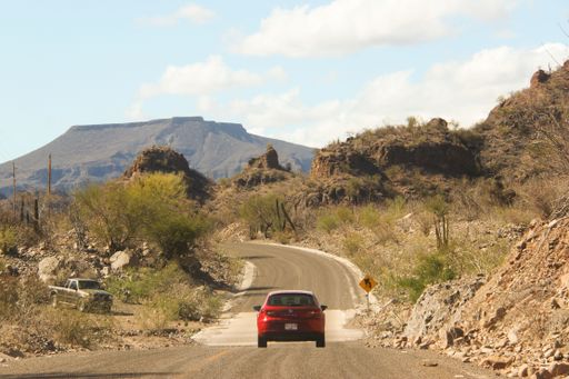A vibrant red car traveling down a winding dirt road surrounded by majestic mountains and sun-bleached desert