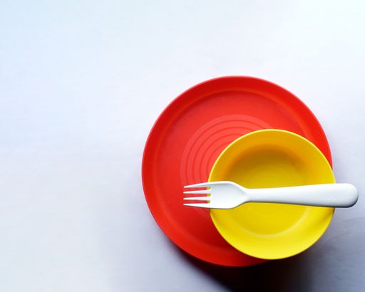 A top view of a red plate, a yellow bowl and a white fork on a white surface with copy space
