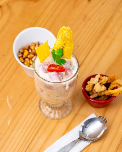 A stainless steel spoon and fork resting on a light-colored wooden table surface near a white ceramic bowl filled with a variety of fresh food items