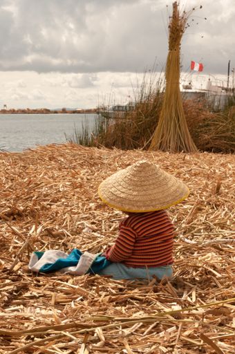 A vertical shot of a child in a wicker conical hat sitting on the coast of the Uros Island, Peru