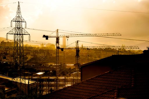 An aerial view of a cityscape at sunset, featuring multiple construction cranes silhouetted against the skyline