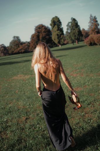 A young woman wearing a black dress stands in a meadow of lush green grass, carrying her shoes in her hand
