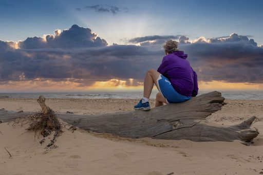 A female adult wearing a purple shirt is seated on a log at the beach