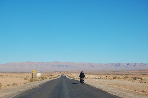 A wide shot of motorcycle riding on a road in the middle of the desert on a sunny day with mountains in the background