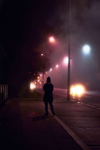 A solitary male figure stands illuminated against a night-time backdrop of a bustling city street