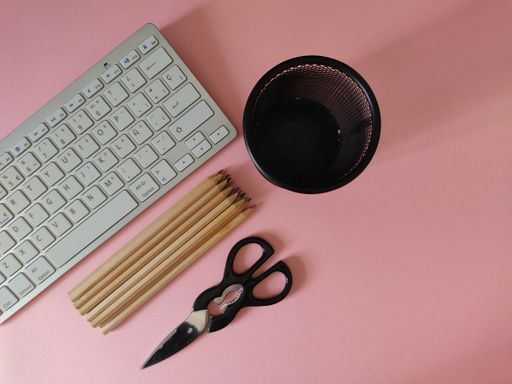 A bunch of colored pencils, a metal pen holder, a white keyboard, on the pink table